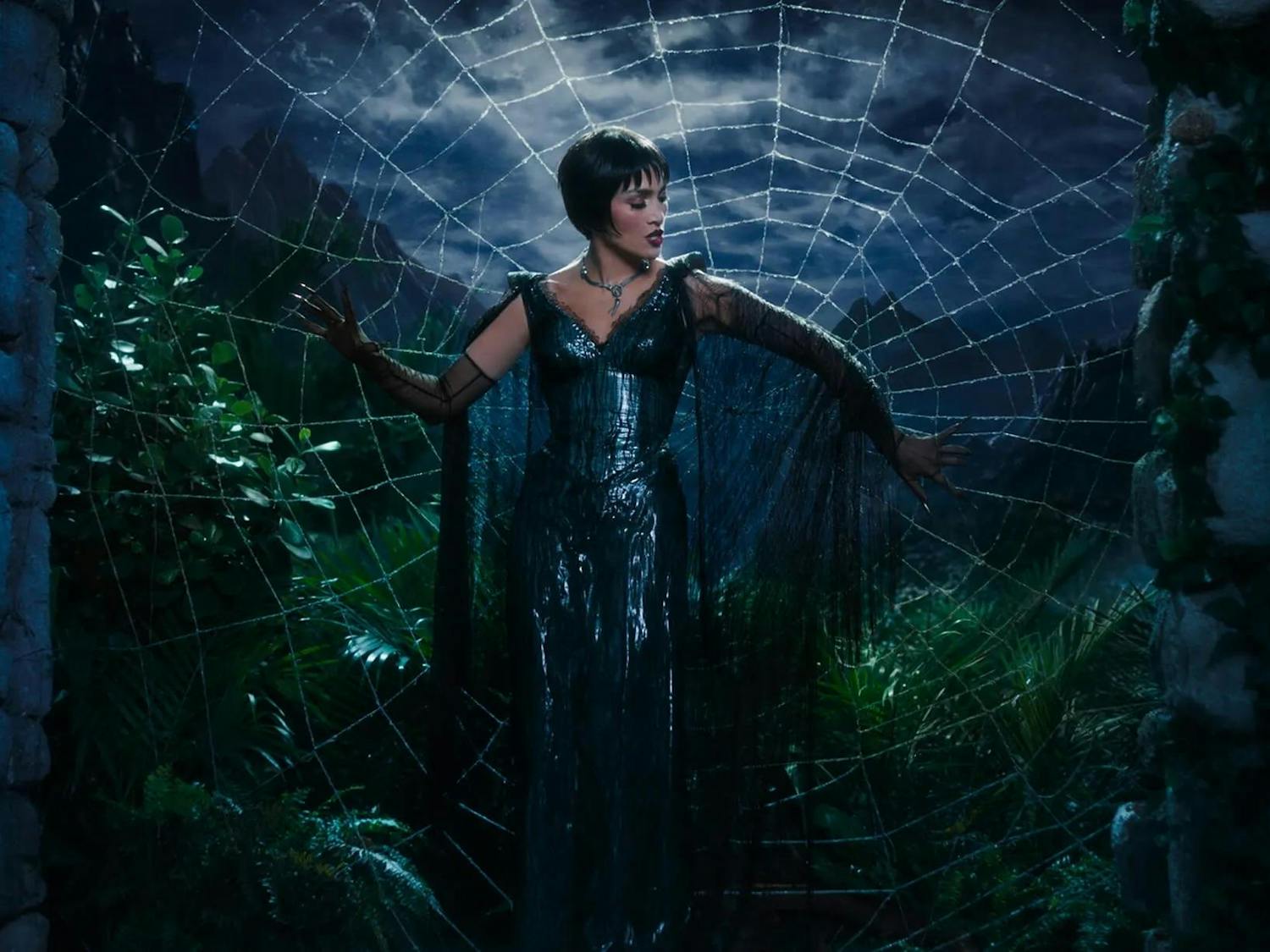 Photo of a woman dressed in a black dress standing in front of a spiderweb with moonlight illuminating the sky in the background.