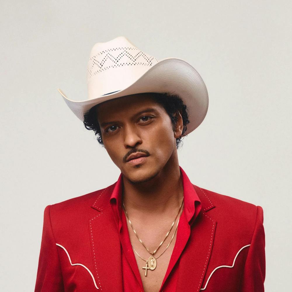 Photo of Bruno Mars wearing a white cowboy hat and a red blazer with two white lines over a red button-up with the top three buttons undone.