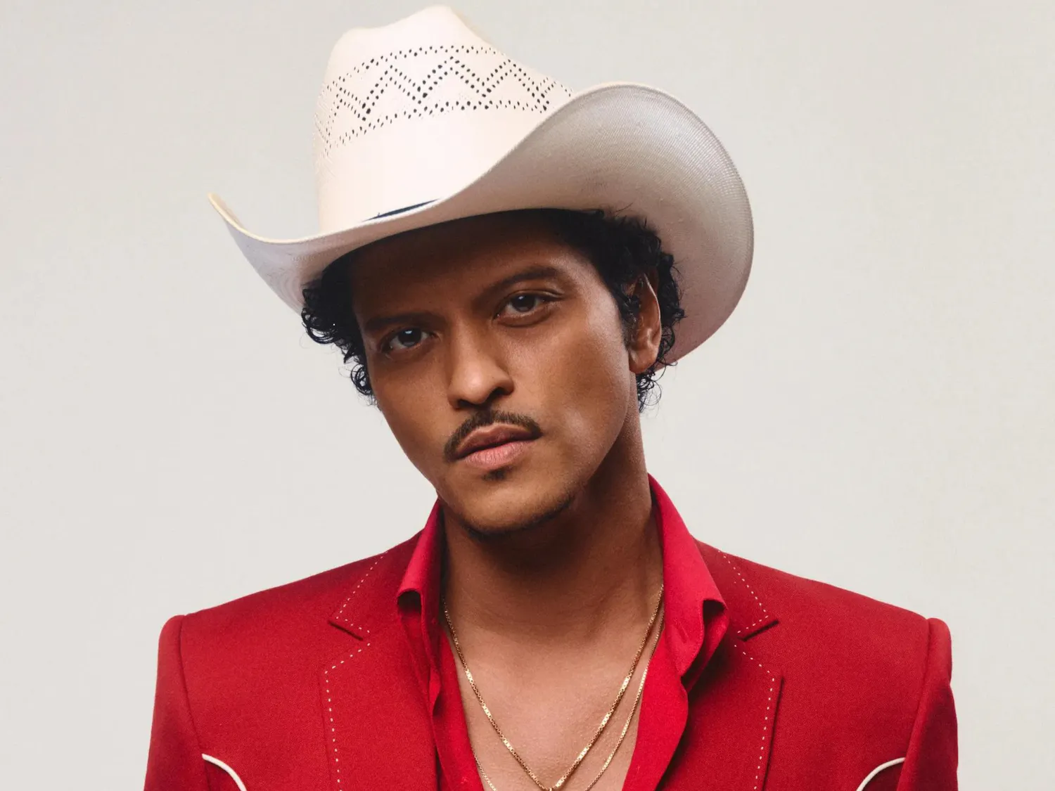 Photo of Bruno Mars wearing a white cowboy hat and a red blazer with two white lines over a red button-up with the top three buttons undone.