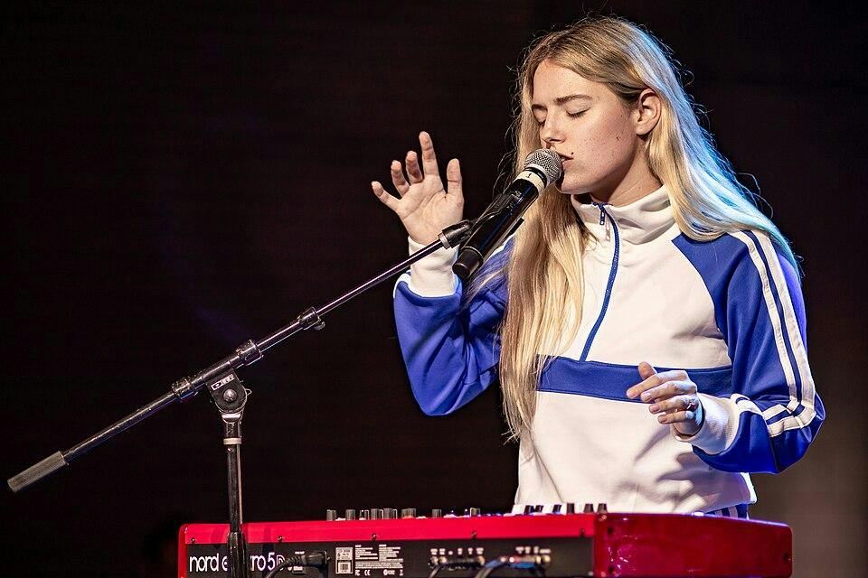 A photo of a blond woman in a white and blue sweatshirt singing in front of a keyboard. 

