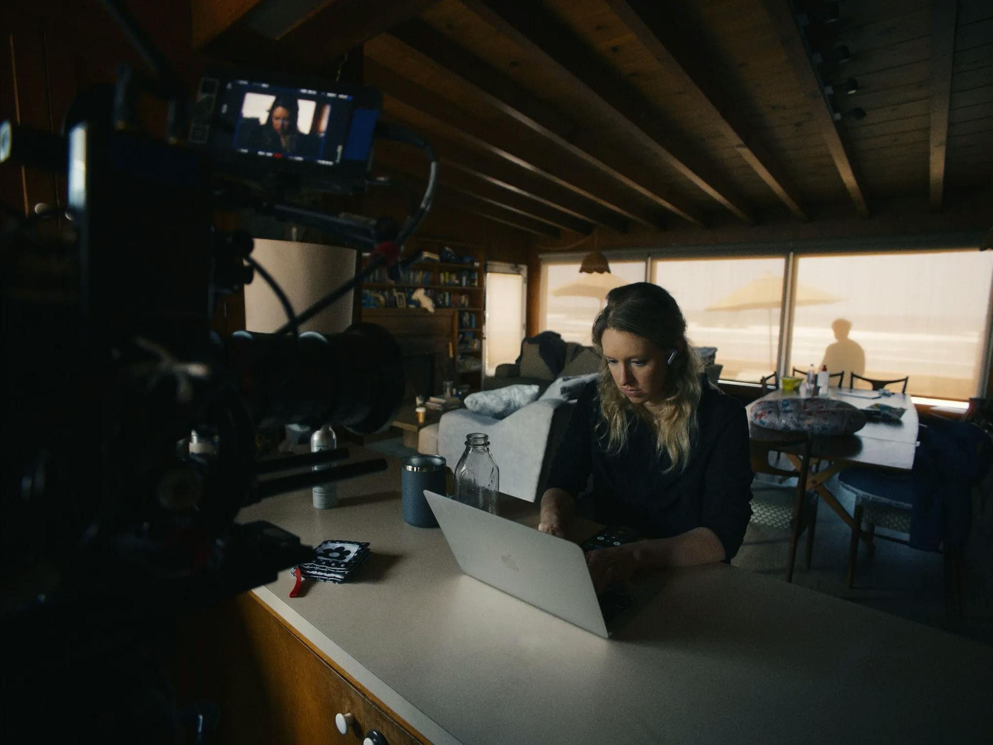 Photo of a woman wearing headphones focused on a silver laptop with a film camera pointed directly at her.