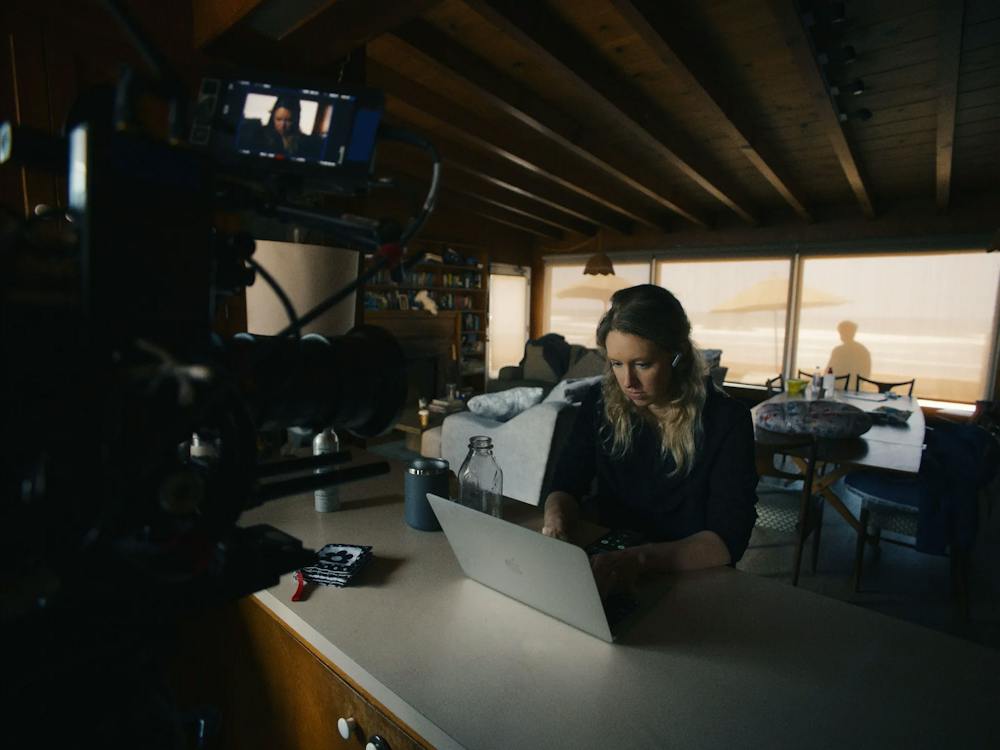 Photo of a woman wearing headphones focused on a silver laptop with a film camera pointed directly at her.