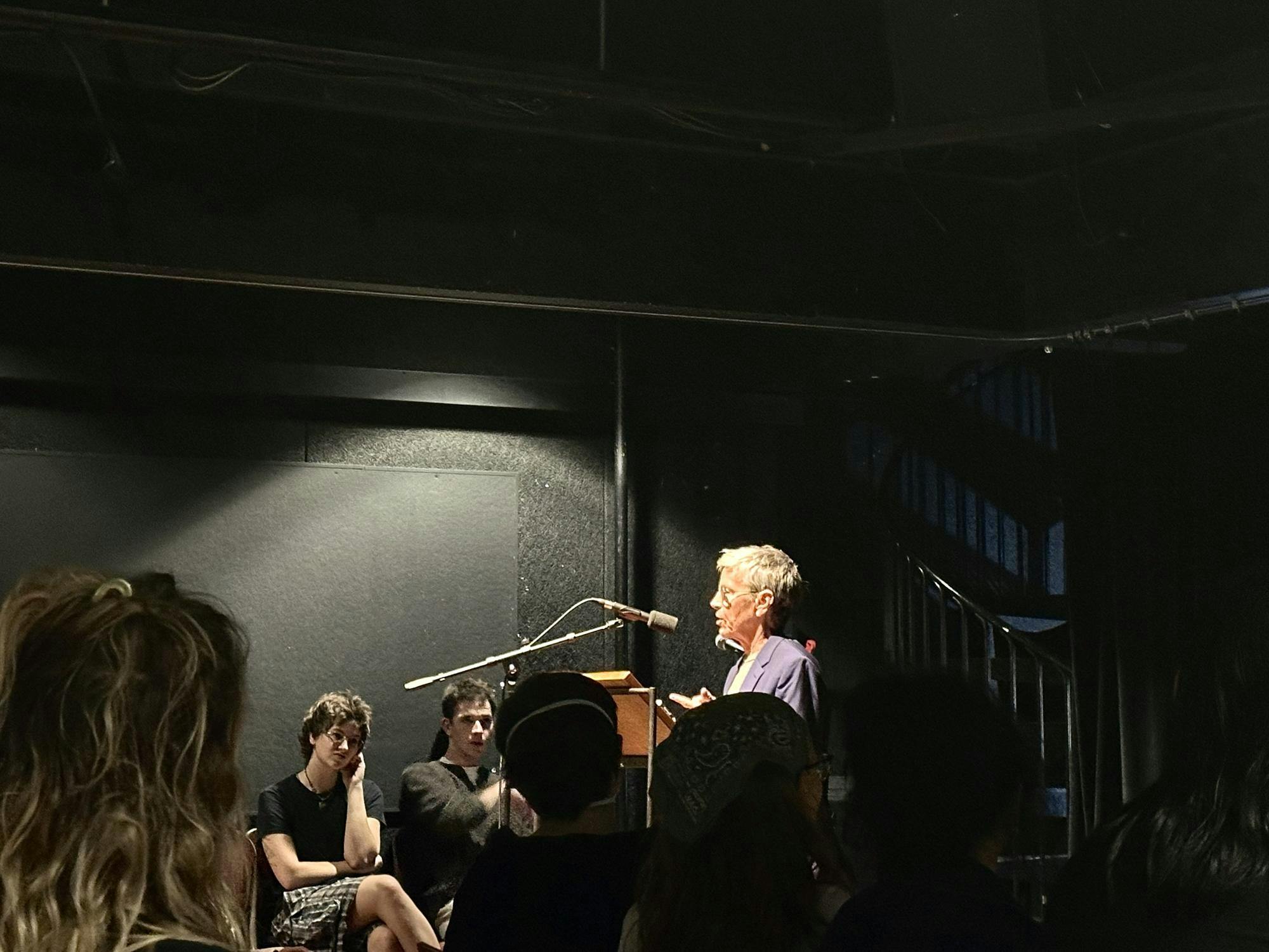 A photo of Eileen Myles standing talking in a theater in a spotlight as an audience listens in the dark.

