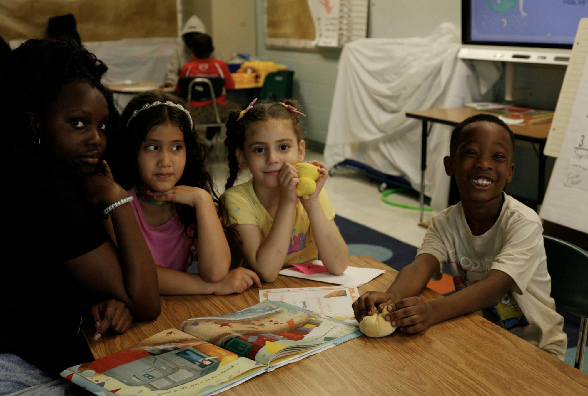 Photo of three kids and their teacher sitting at a table with an open picture book between them.