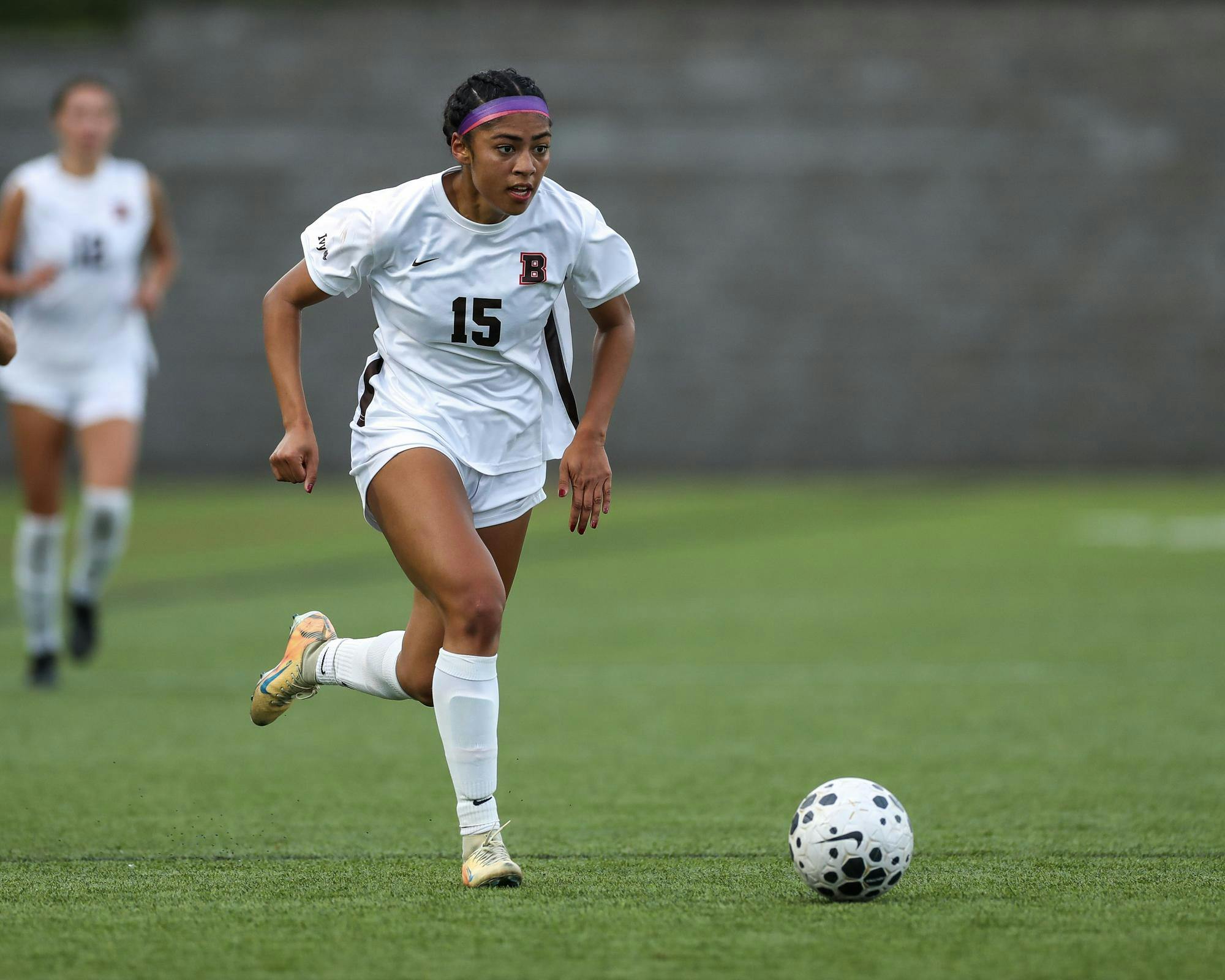 Photo of a Brown soccer player wearing a white uniform running on the field and dribbling a soccer ball. 
