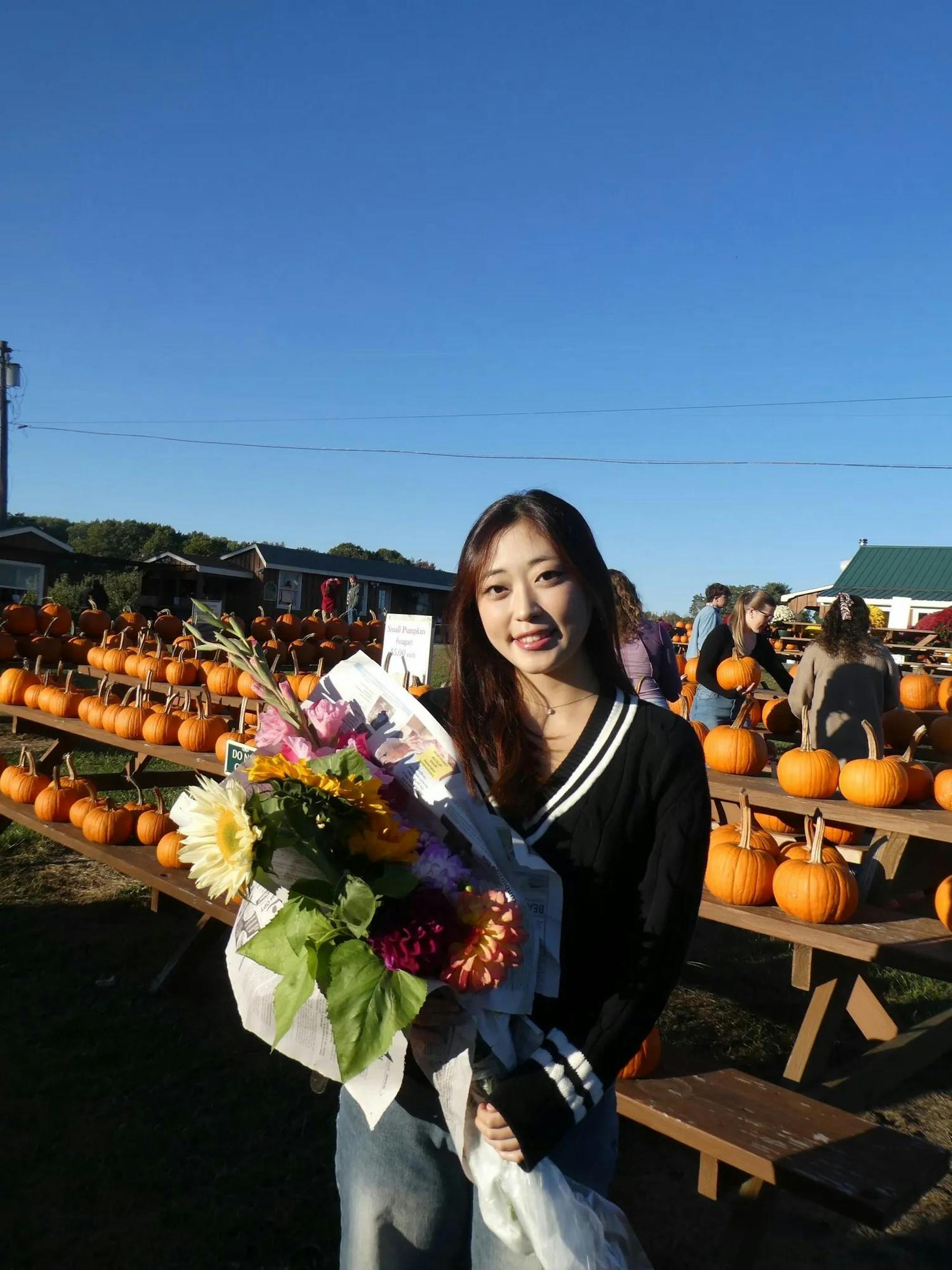 Photo of a girl wearing a black and white cardigan and jeans holding a large bouquet of flowers with pumpkins lining picnic tables in the background.