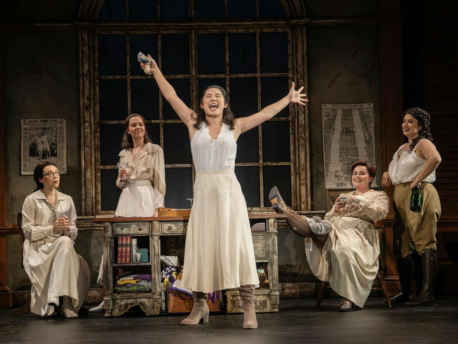 A woman on stage wearing early 19th-century casual garments holds her arms out in passion while mid-song. Four other women behind her watch her perform fondly.