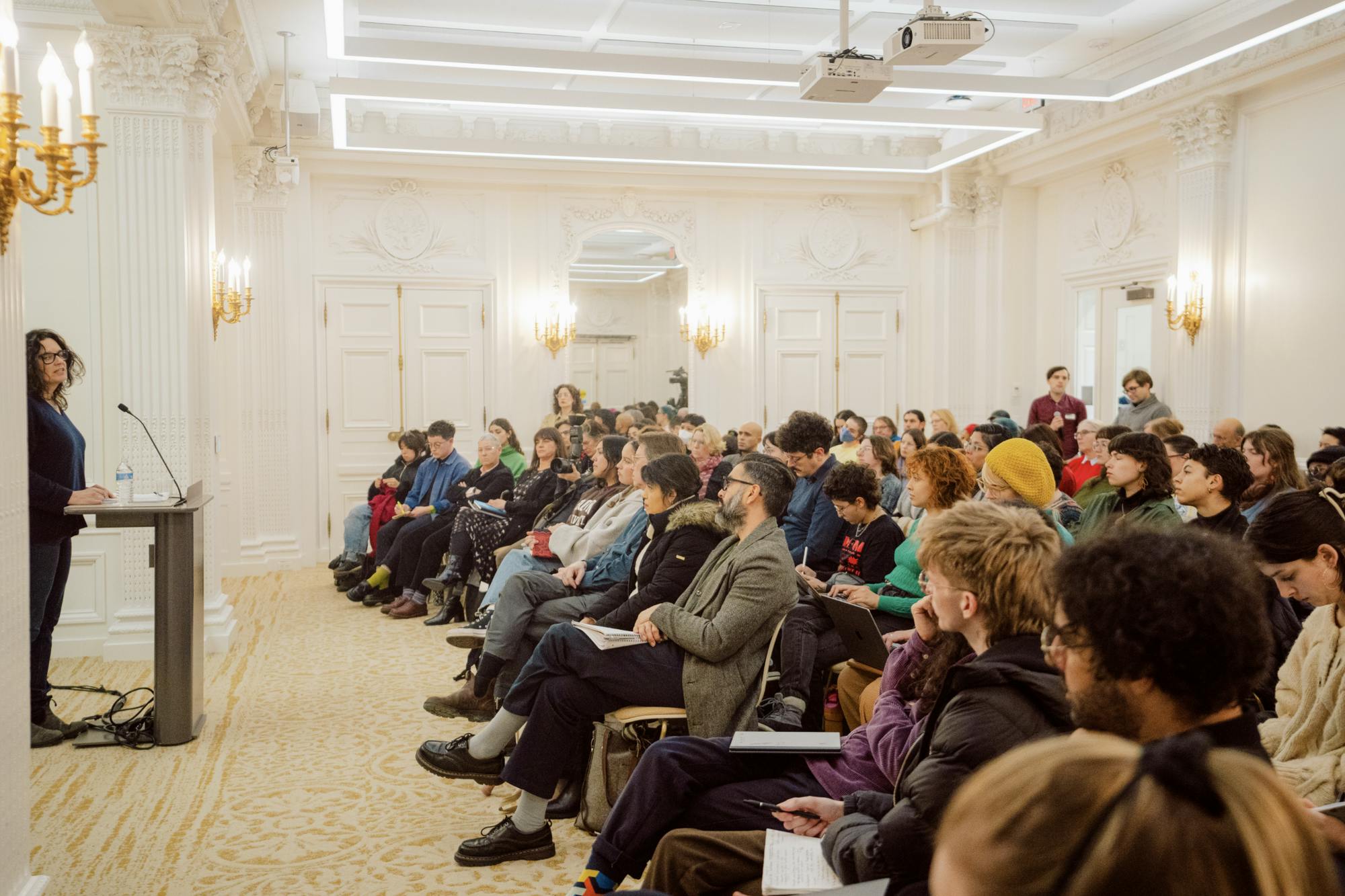 Leanne Betasamosake Simpson, the event's lecturer, stands in a bright room with white walls and golden lamps as she speaks to the crowd of attendees.