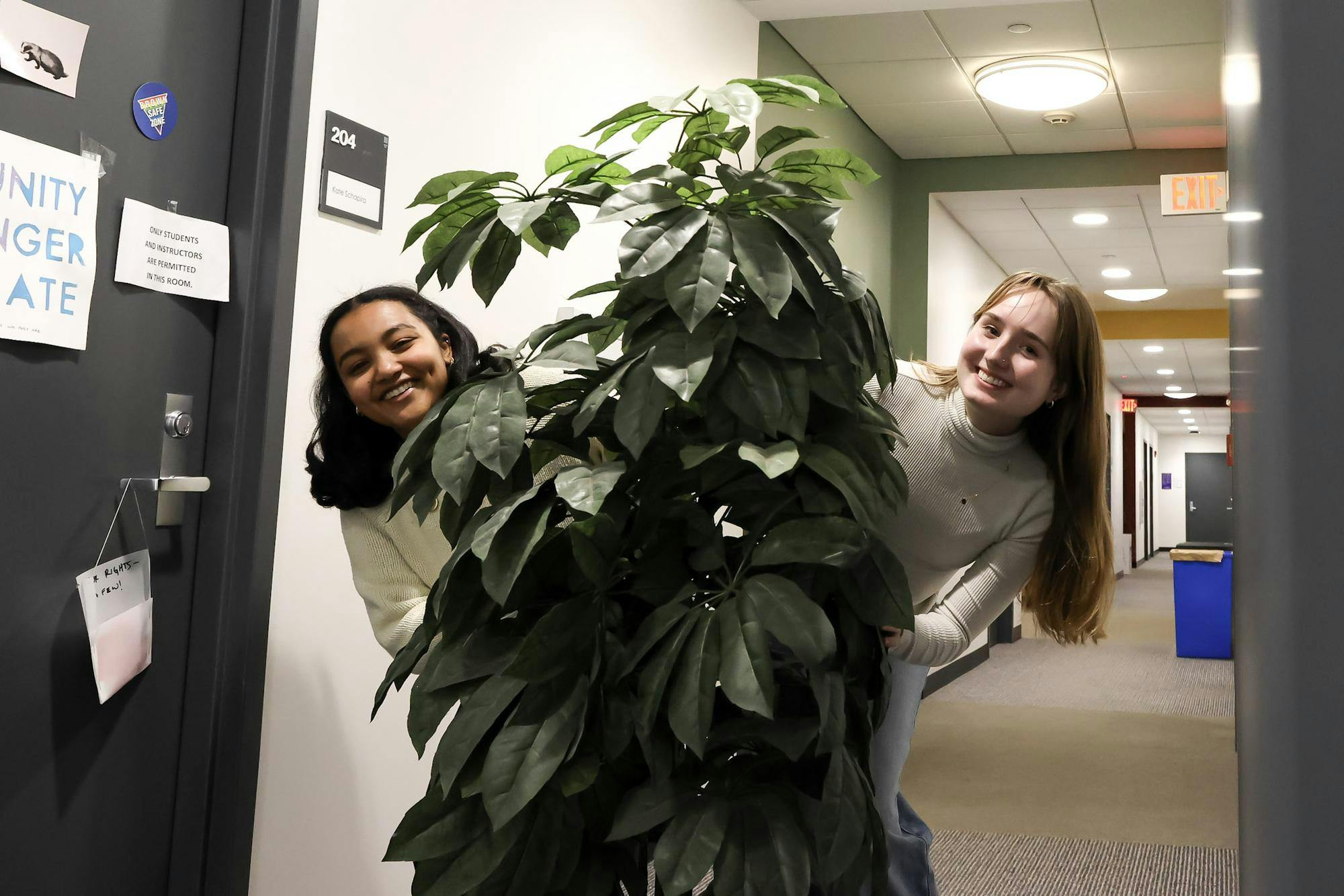 A photo of Lydia Fantaye (left) and Grace Gongoleski (right) peering out the sides of a plant in a hallway.