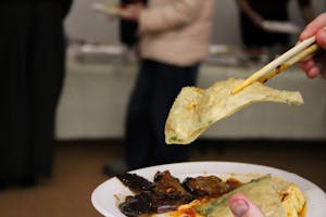 Photo of a pair of chopsticks holding a scallion pancake with an assortment of food on a plate held beneath.

