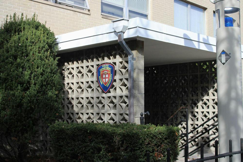 Photo of the outside entrance of the Brown University Department of Public Safety Building: A concrete building with a blue shield logo on the side with the text “Police Department Brown University” surrounded by bushes.