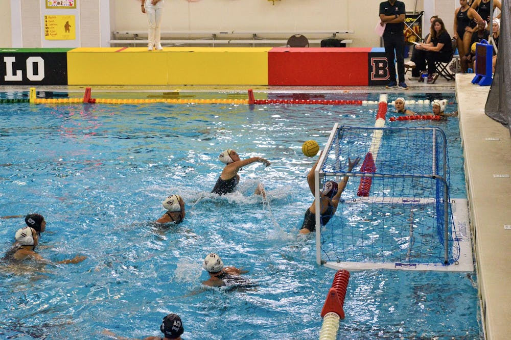 A photo showing a water polo game in the pool where a player is shooting a ball at a goal that is being blocked by a Brown University goalie, with other teammates surrounding them.