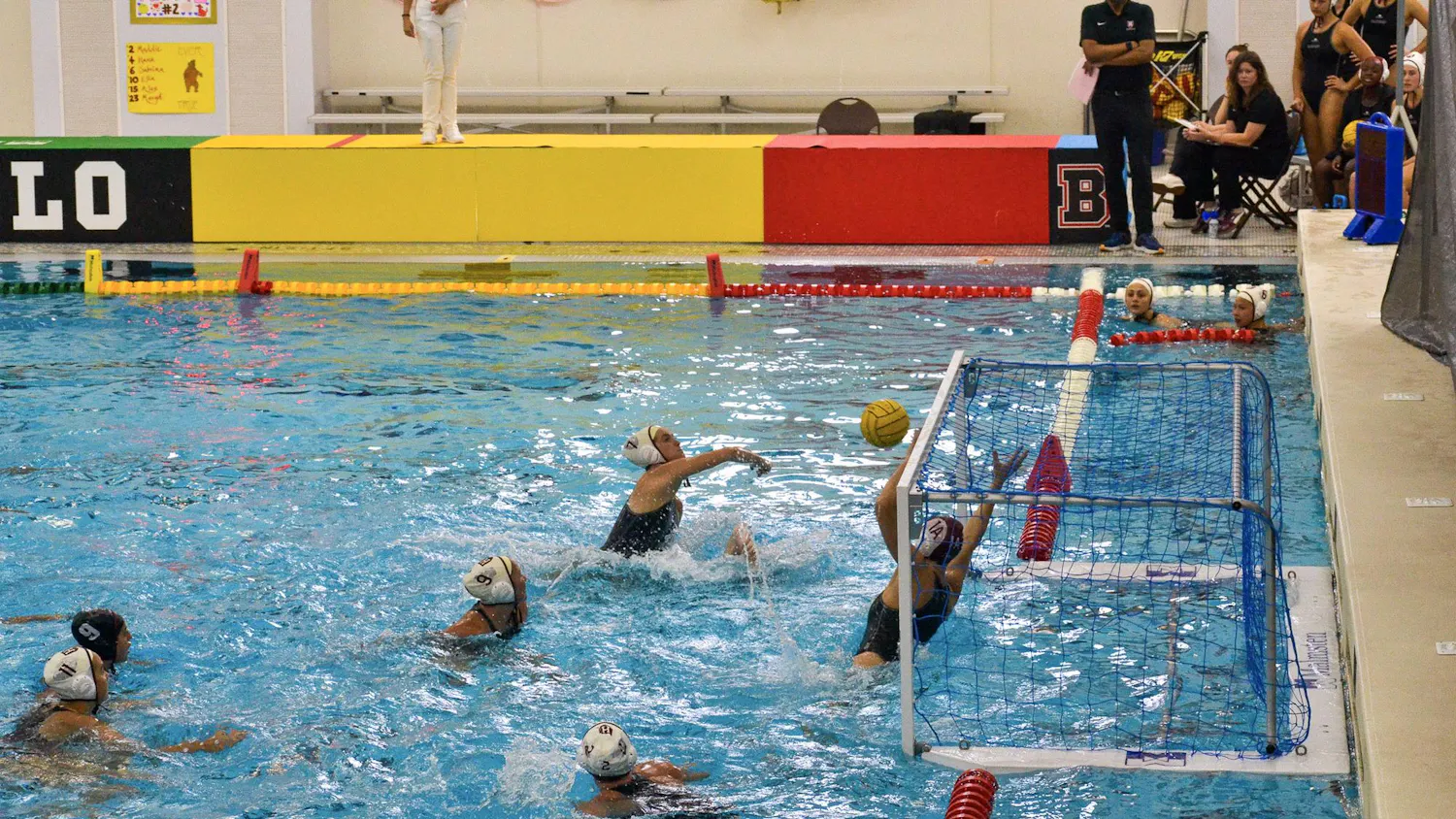 A photo showing a water polo game in the pool where a player is shooting a ball at a goal that is being blocked by a Brown University goalie, with other teammates surrounding them.