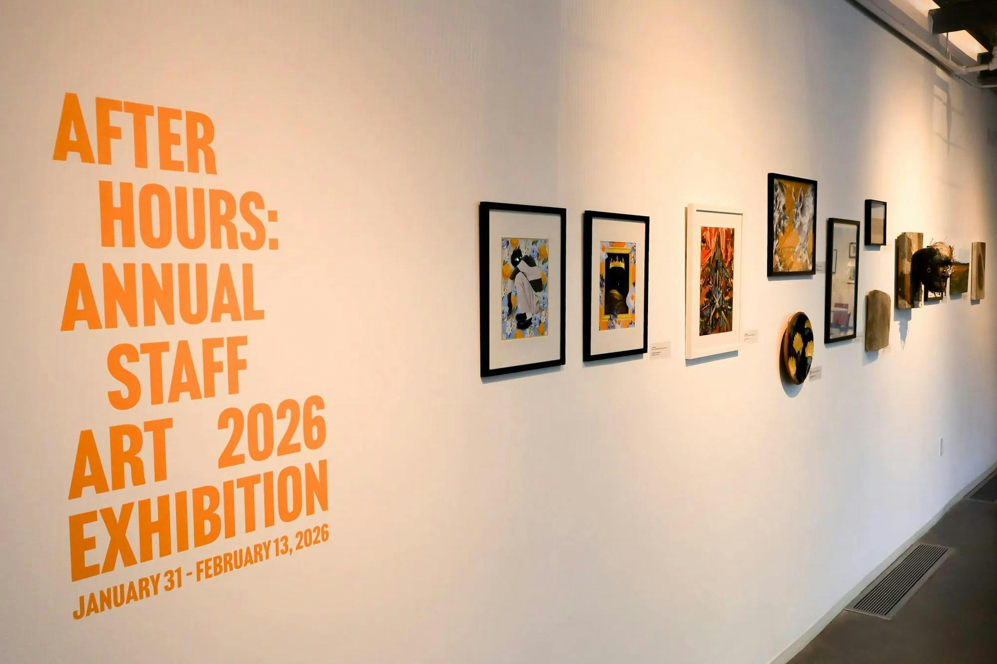 Photo of a white wall with orange print that states “AFTER HOURS: ANNUAL STAFF ART 2026 EXHIBITION / JANUARY 31 - FEBRUARY 13, 2026” alongside a row of paintings and other artwork.

