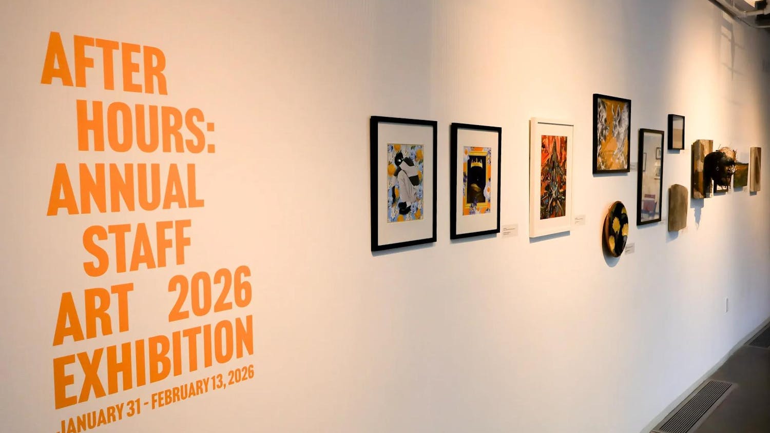 Photo of a white wall with orange print that states “AFTER HOURS: ANNUAL STAFF ART 2026 EXHIBITION / JANUARY 31 - FEBRUARY 13, 2026” alongside a row of paintings and other artwork.