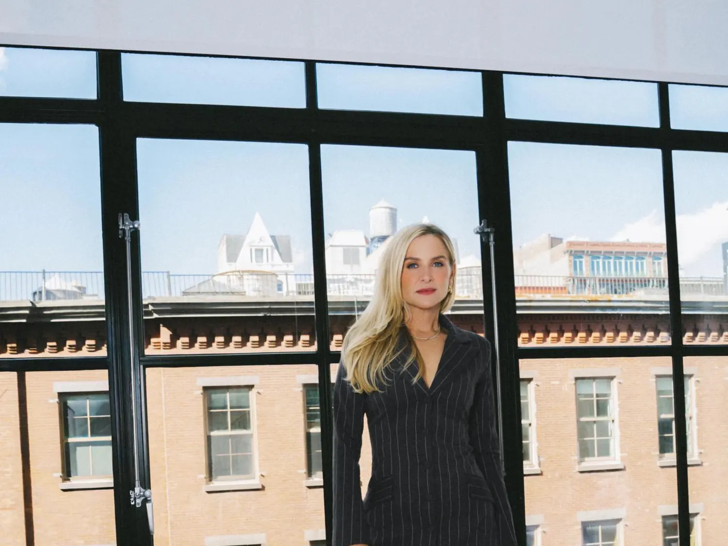 Image of Jessica Capshaw in a black striped dress and blue heels standing against the windows, featuring multiple buildings in the background.