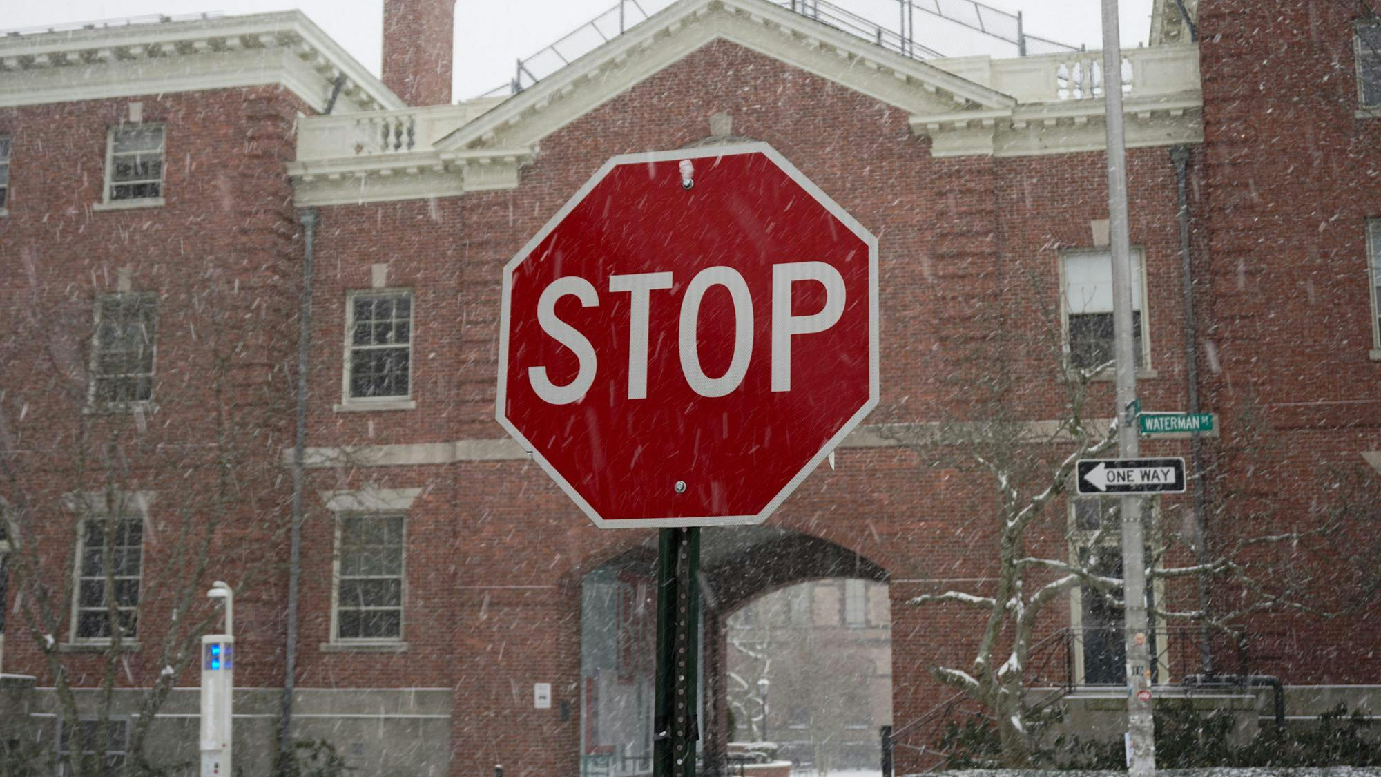 Photo of a stop sign with snow falling around it. The sign is in front of a red brick building.

