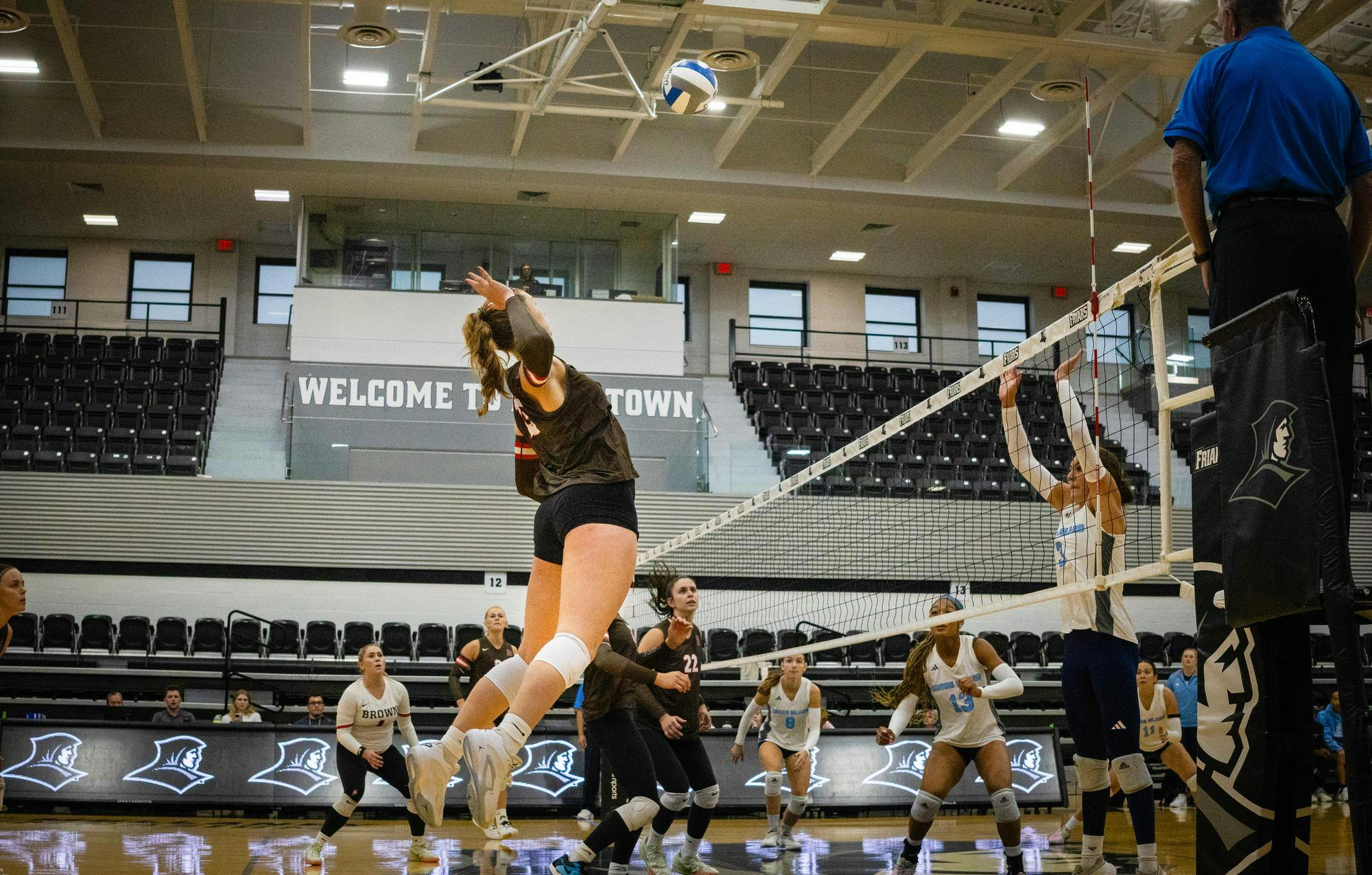 A photo of volleyball players, some dressed in brown, others dressed in white. In the center a player prepares to spike a ball. 