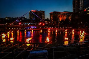 A circular lake with stakes poking out that are lit on fire surrounding the body of water. It is dark outside and there are buildings behind the body of water illuminated by the orange glow.