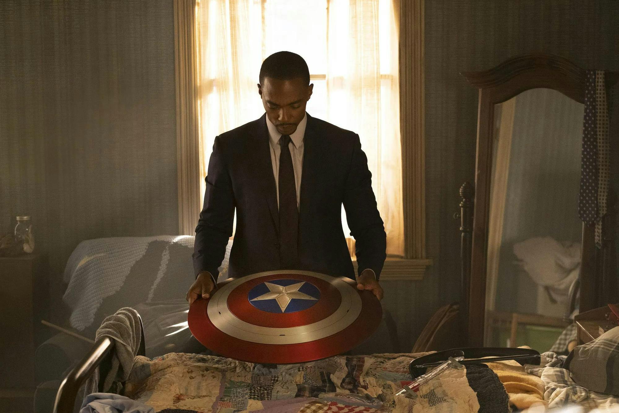 Captain America, played by Anthony Mackie, stands in a black suit in a softly lit bedroom holding the red, white and blue Captain America shield.