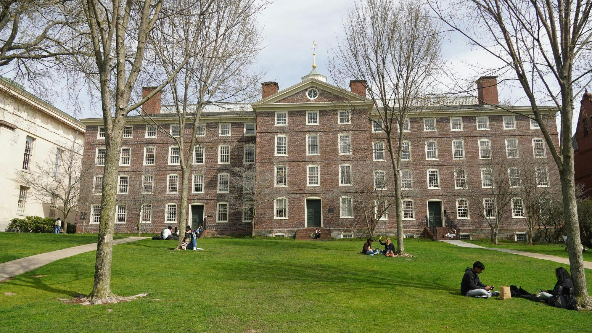 A photo of University Hall from the Quiet Green on a sunny day. Various groups of students are sitting on the green.