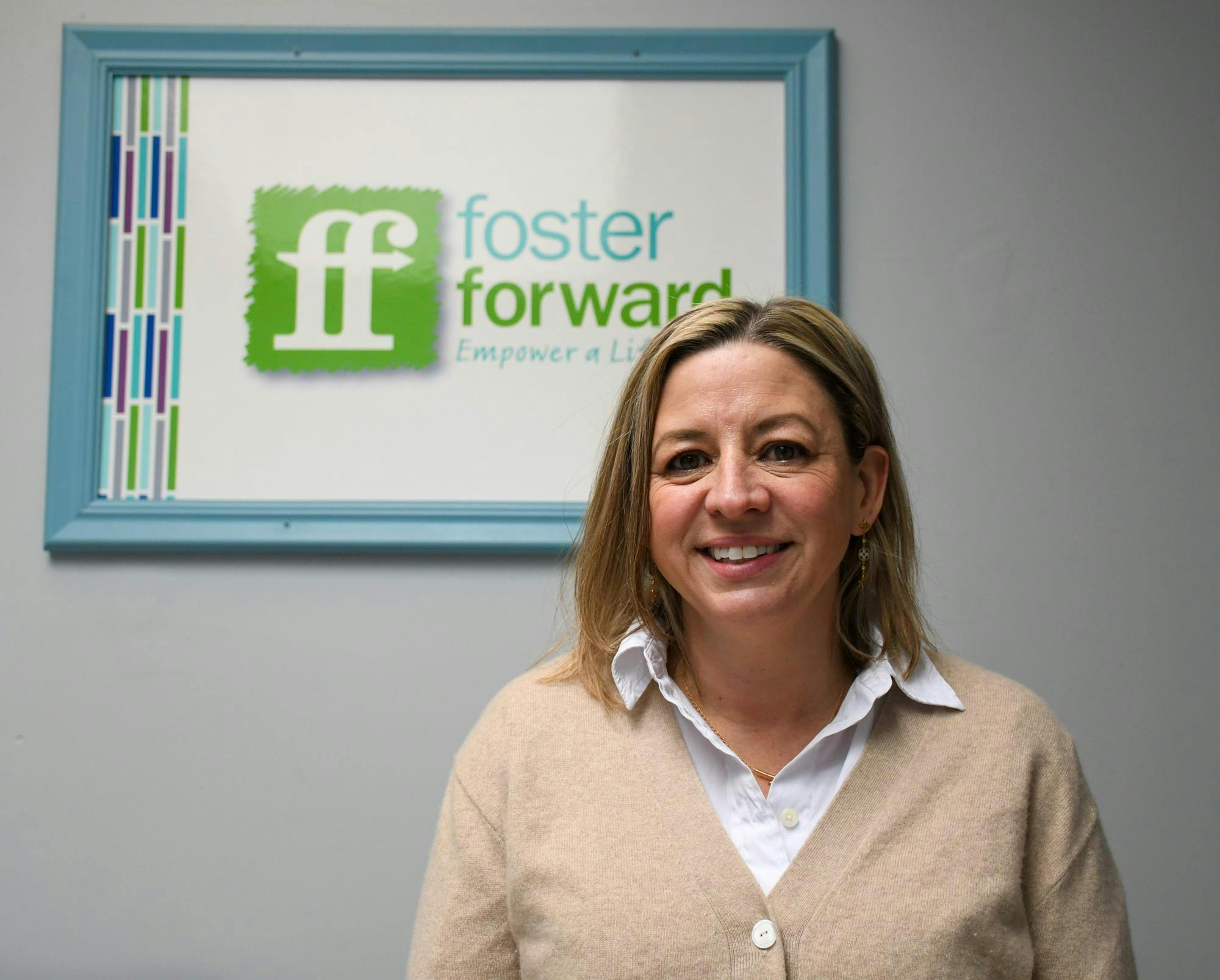 Lisa Guillette stands in front of a green and blue foster forward sign. She has blonde hair and is wearing a tan cardigan over a white collared shirt. 