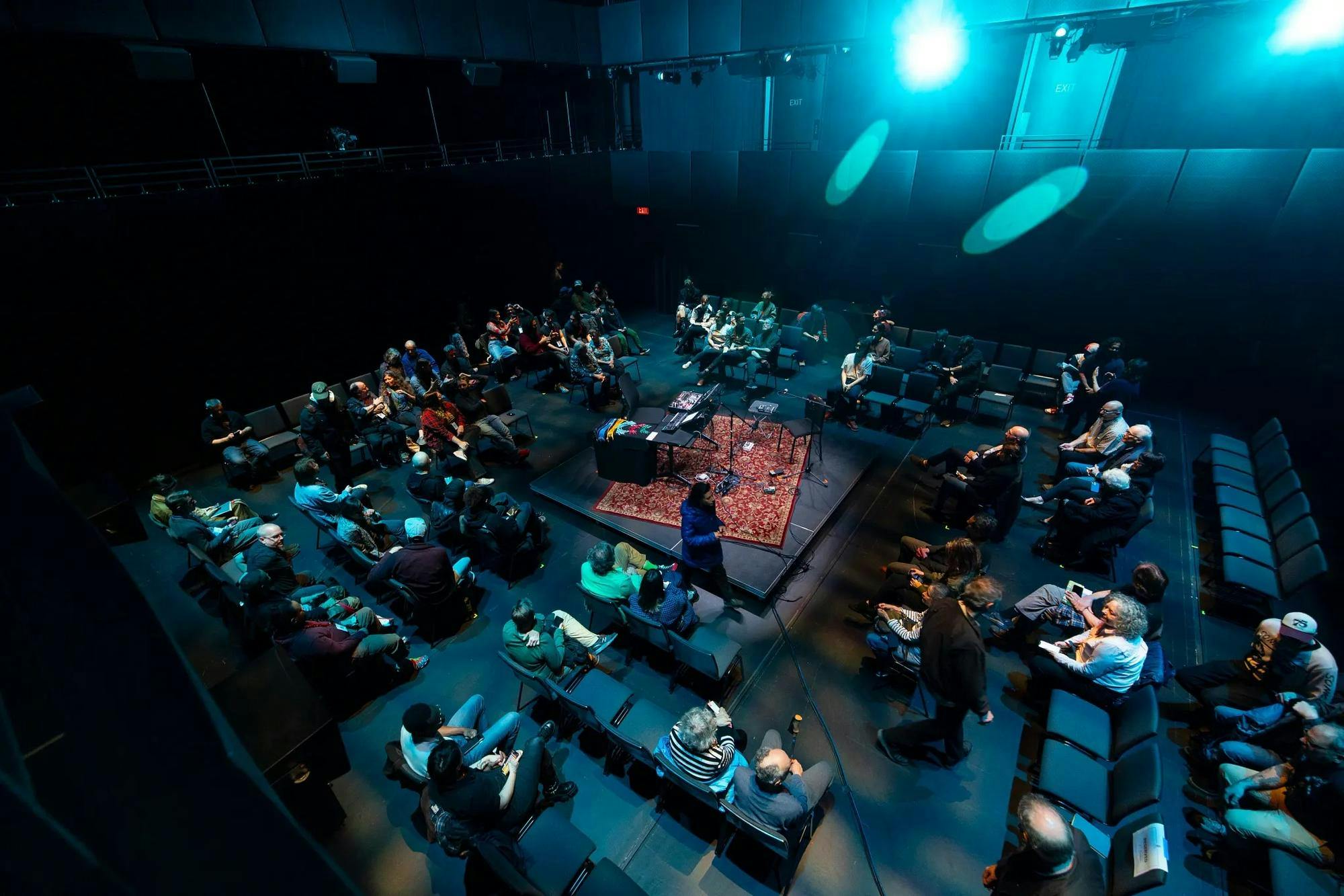 People in a blackbox theater with a rug in the middle of the room with someone kneeling on it.