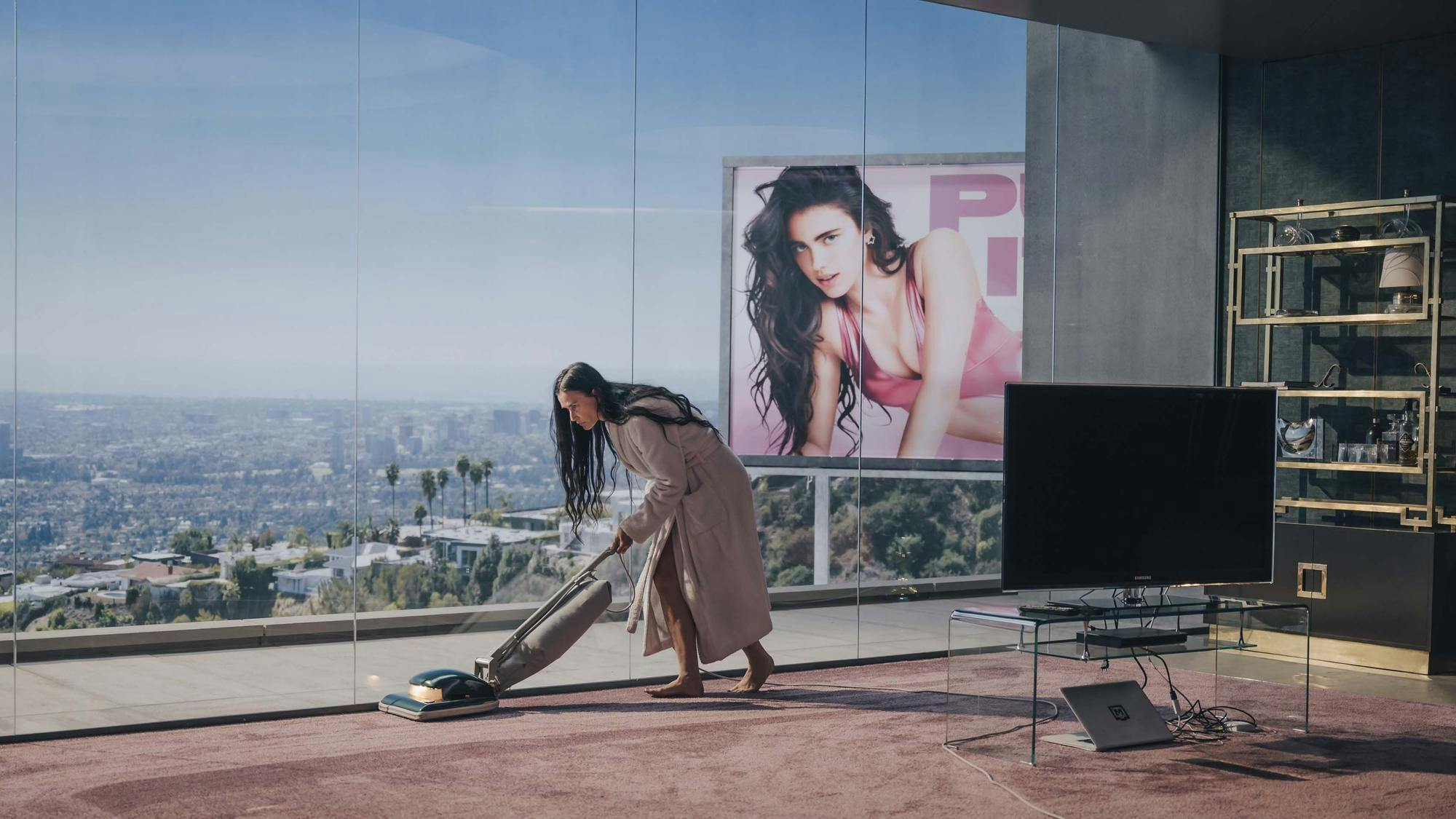 A woman in an LA apartment with floor-to-ceiling windows vacuums a pink carpet. In the background is a billboard with a woman on it. 
