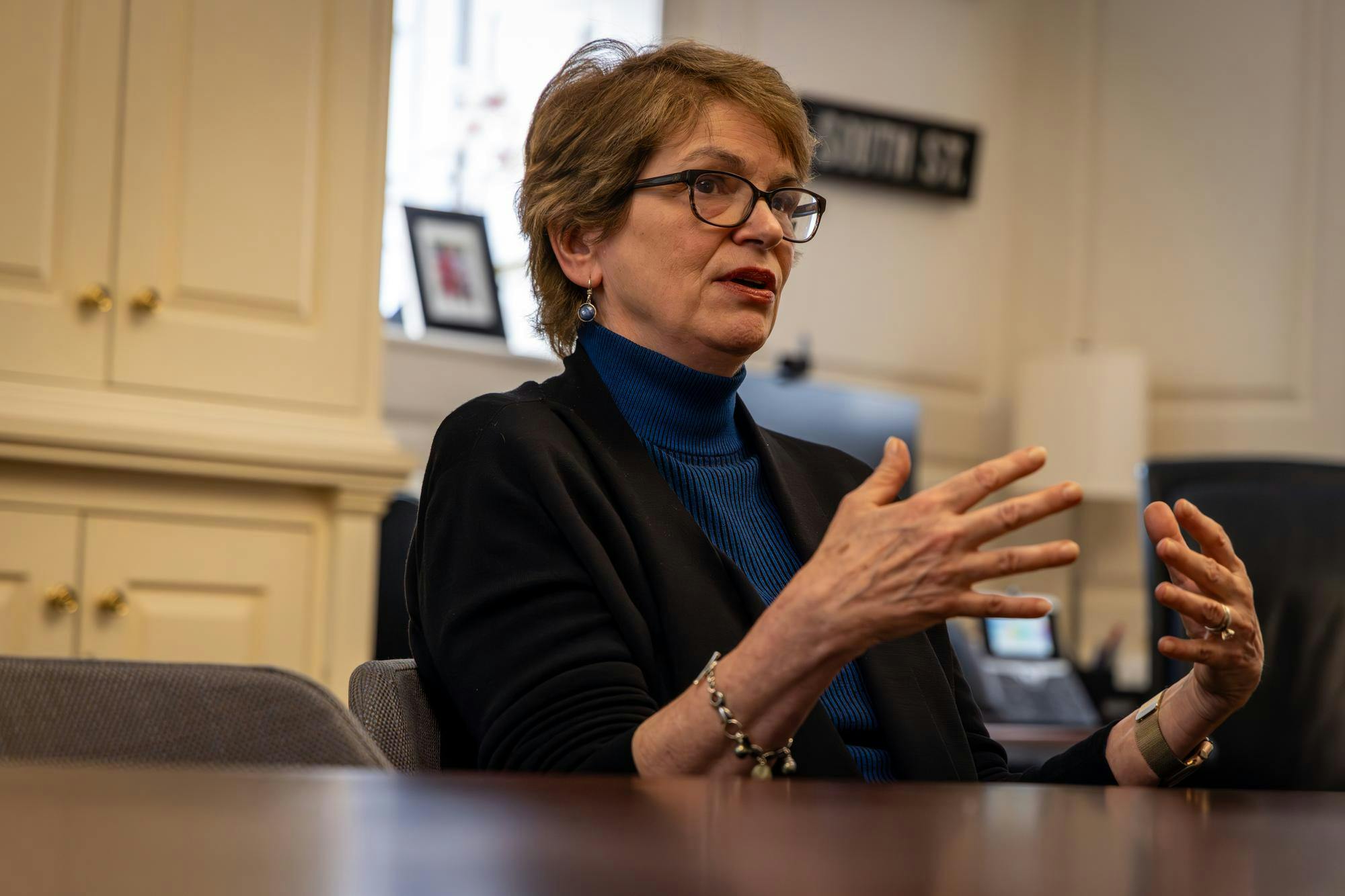 A photo of Christina Paxson wearing a black jacket over a blue turtleneck. She is sitting at a wooden table in a formal office.