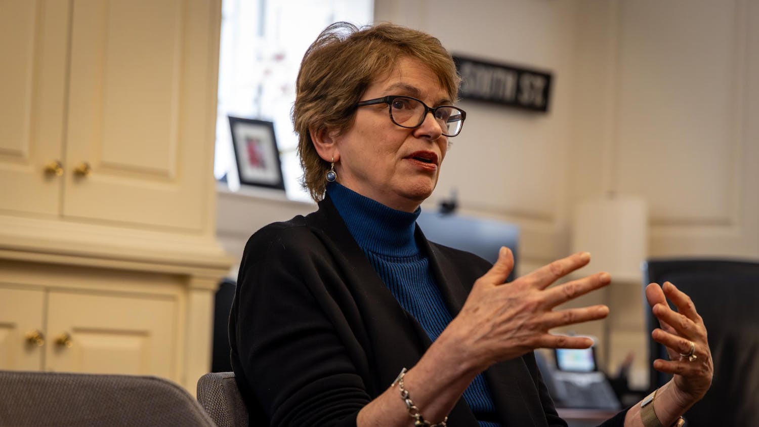 A photo of Christina Paxson wearing a black jacket over a blue turtleneck. She is sitting at a wooden table in a formal office.
