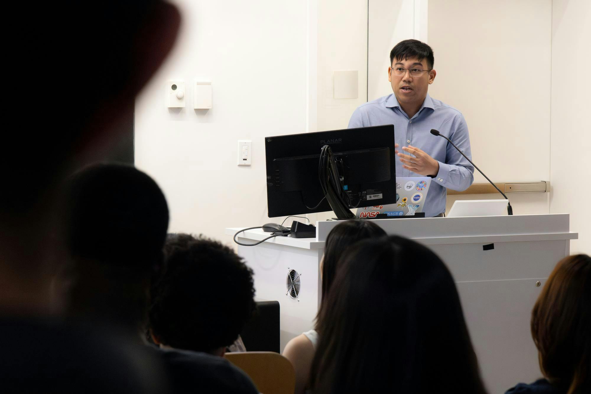 Photo of a man wearing a blue collared shirt standing at a white podium speaking to a crowd.