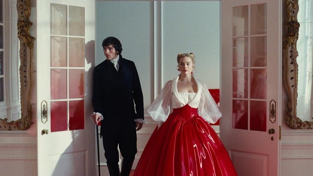 Shot from “Wuthering Heights” (2026) featuring Heathcliff (Jacob Elordi) and Catherine Earnshaw (Margot Robbie) walking through a door frame. Robbie wears a red and white gown and Elordi wears a black suit and holds a cane. 