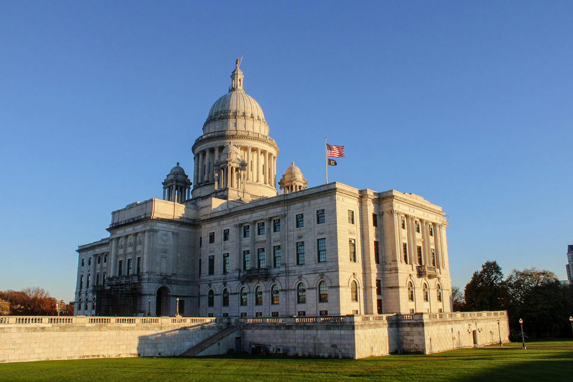 Picture of the Rhode Island State House from afar. An American flag waves in the wind. Overhead, the sky is blue and cloudless.