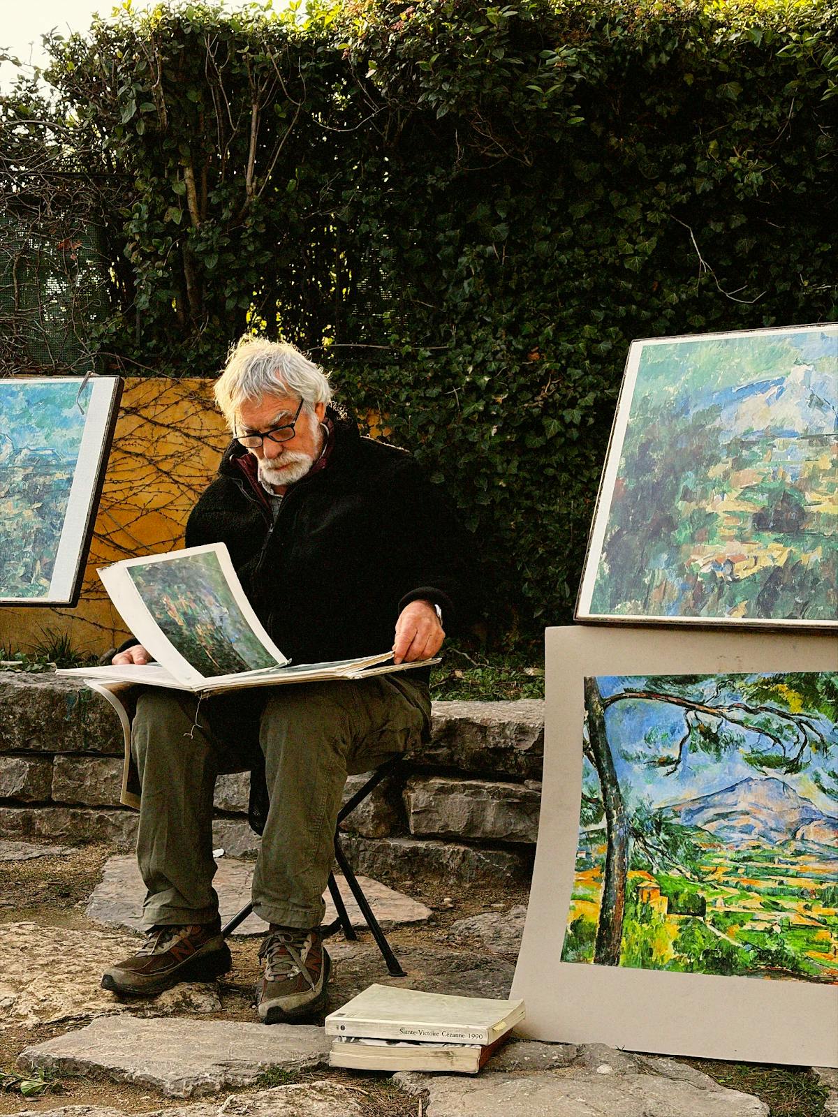 Photo of a man sitting on a stool flipping through artwork, surrounded by paintings in front of a bush.