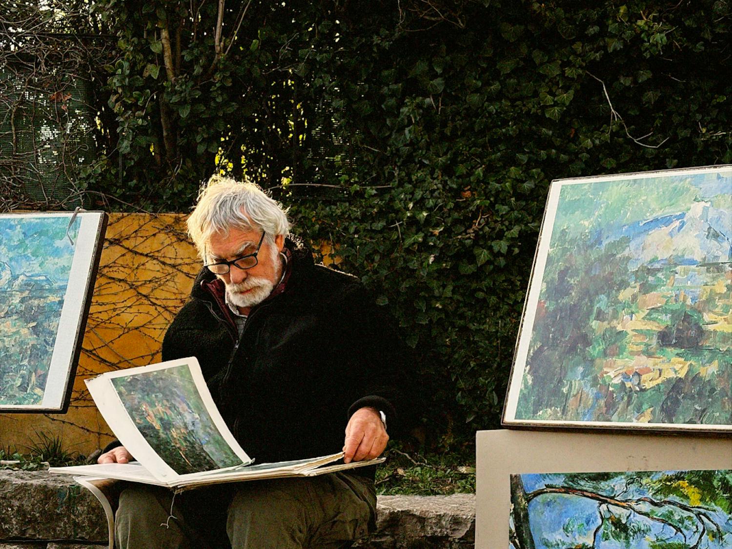Photo of a man sitting on a stool flipping through artwork, surrounded by paintings in front of a bush.