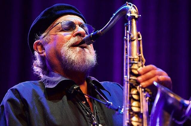 Joe Lovano, wearing a black collared shirt and a cap, pictured playing the saxophone. 