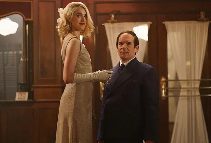 A blonde-haired woman in a gold dress stands next to a seated man in a blue suit in a room adorned with dark wood. 