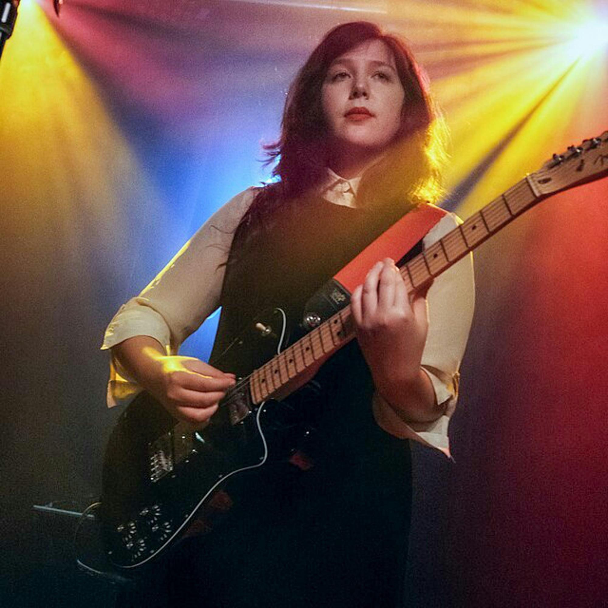 Lucy Dacus wearing a white collared shirt with a black sweater playing a guitar surrounded by yellow and red lights.