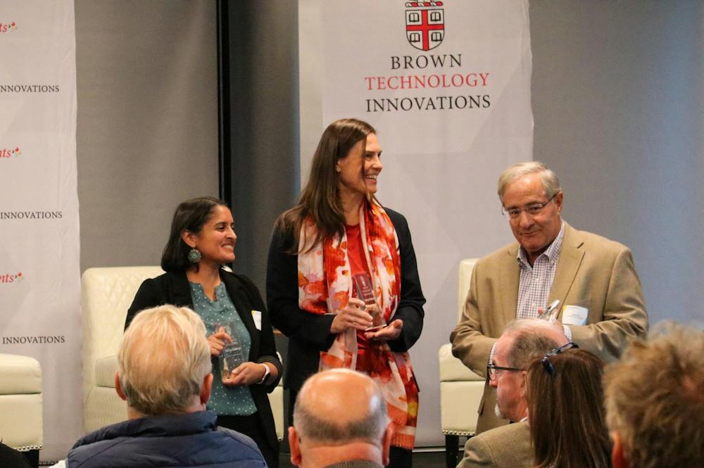 A photo of a woman in a blue top holding a glass award standing next to another woman in a pink top holding a glass award standing next to a man in a beige blazer standing in front of an audience. Behind them is a sign that has the words “Brown Technology Innovations” on it.