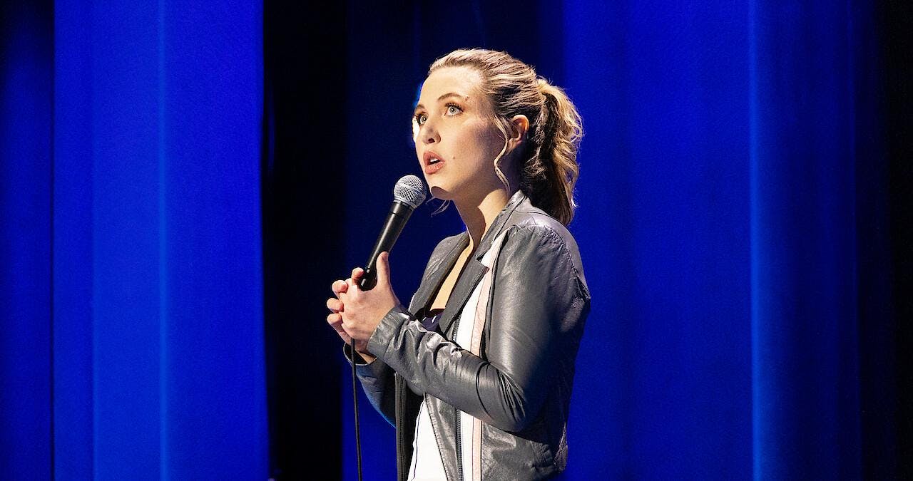 Taylor Tomlinson wears a grey leather jacket and stands in front of sky blue curtains while looking up and holding a microphone.