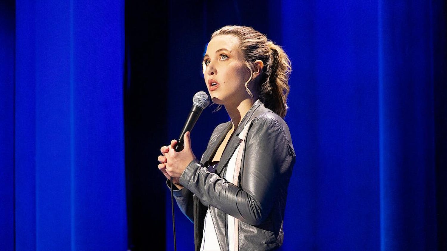 Taylor Tomlinson wears a grey leather jacket and stands in front of sky blue curtains while looking up and holding a microphone.