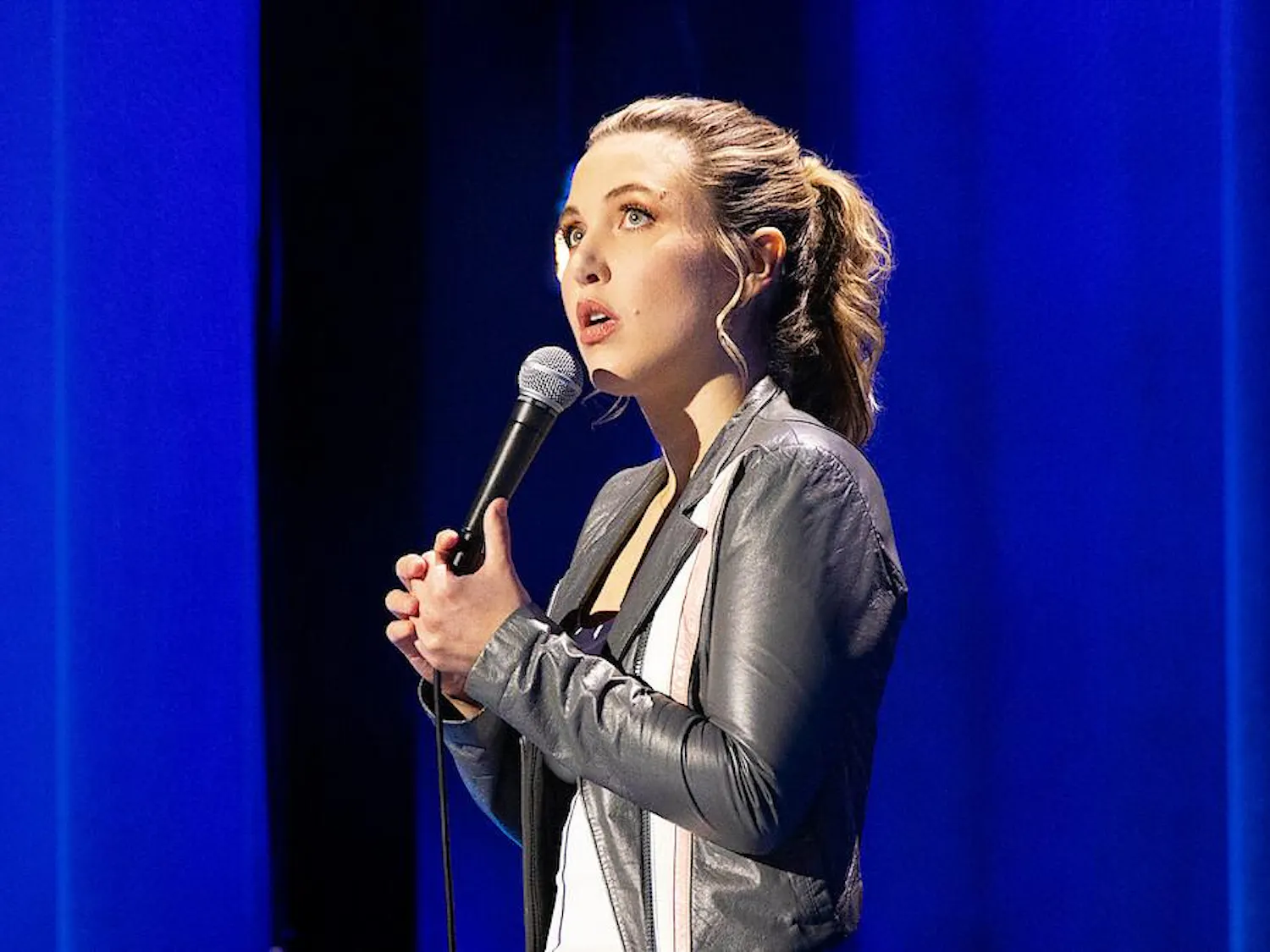 Taylor Tomlinson wears a grey leather jacket and stands in front of sky blue curtains while looking up and holding a microphone.