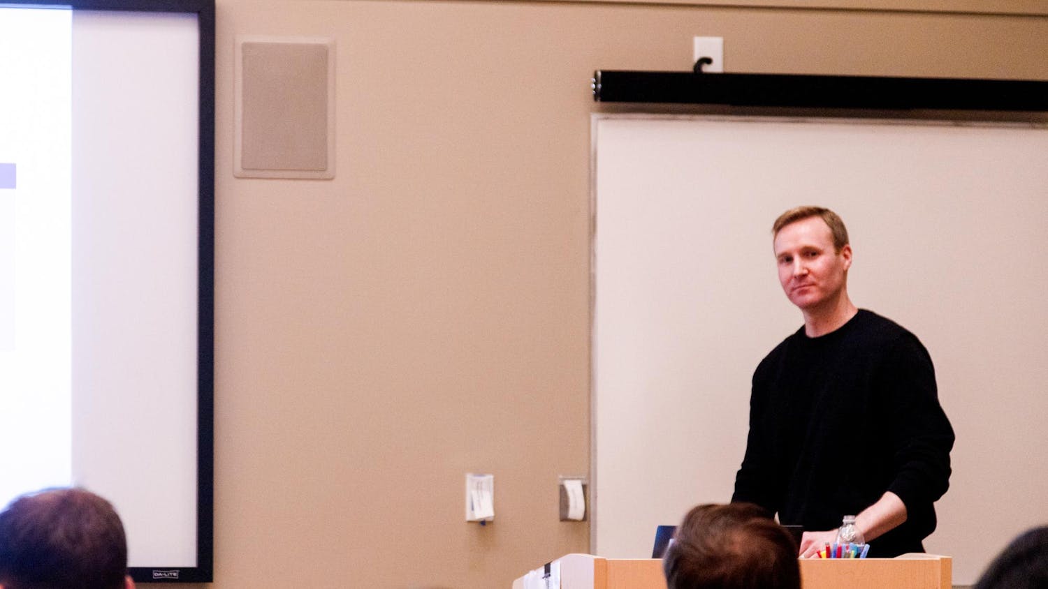 Photo of Will Dobbie at a wooden stand giving a lecture to a group of students.