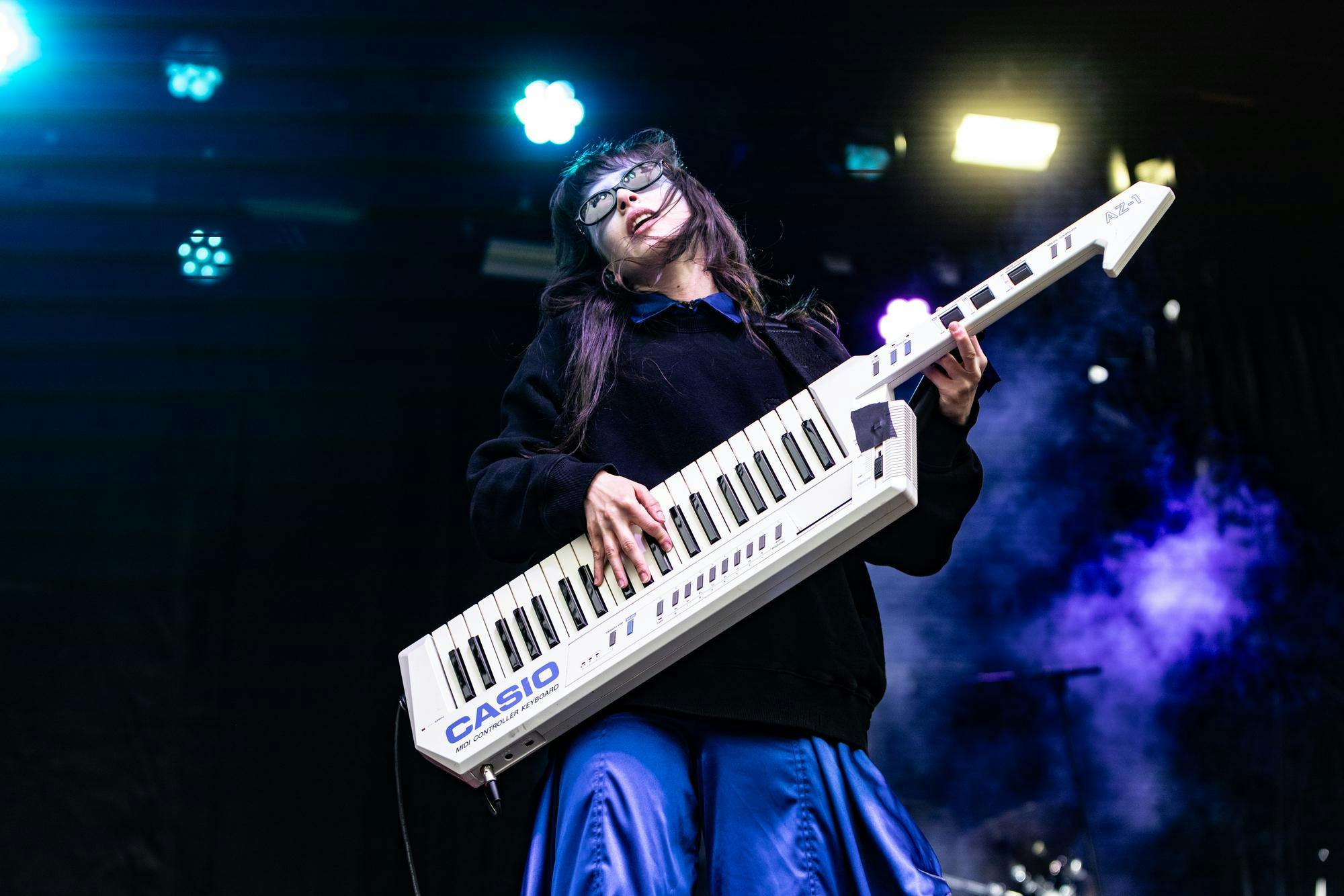 Photo of Mica Tenenbaum, a member of the music duo Magdalena Bay, playing a keytar with colorful lights behind her.