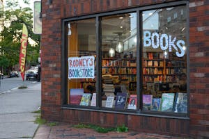 A photo of the brick exterior of Rodeny’s bookstore. Bookshelves are visible through a large window.