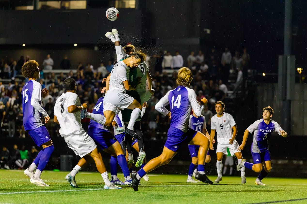 A photo of soccer players, some dressed in purple, others dressed in white. In the center, multiple players crash into each other.