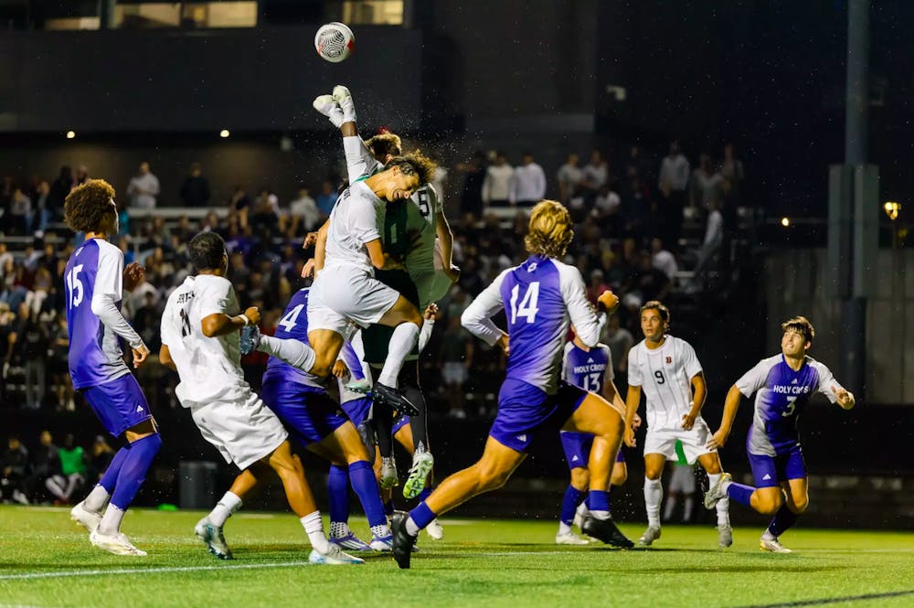 A photo of soccer players, some dressed in purple, others dressed in white. In the center, multiple players crash into each other.