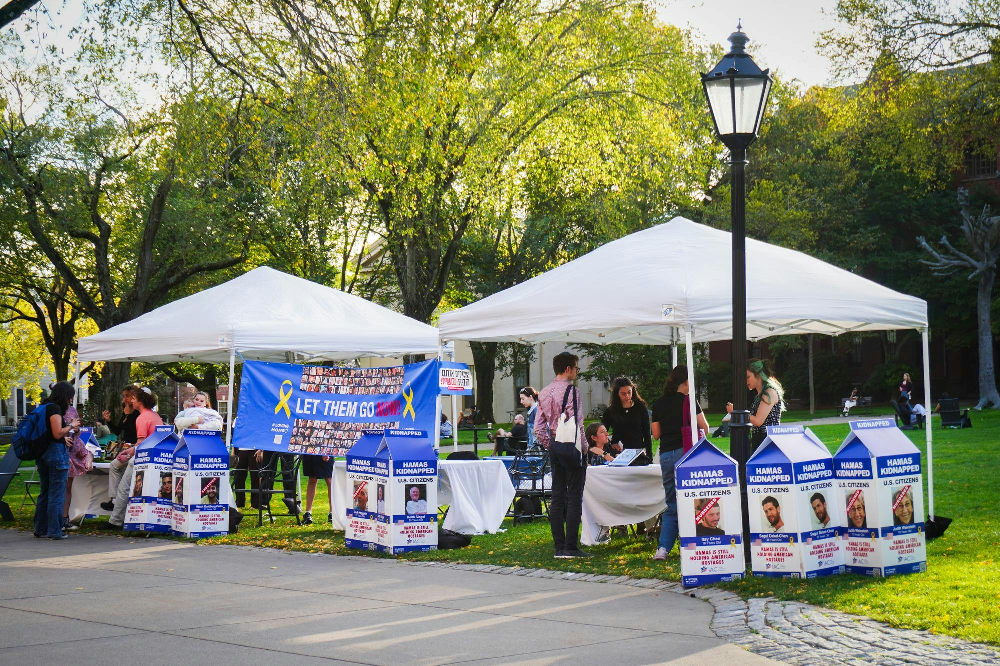 Blue and white display on a green field titled with a blue banner saying "Let Them Go Now". The display is made up of two white tents and tables. In front of the tables are multiple large cardboard signs emulating milk cartons with kidnapping victims. 