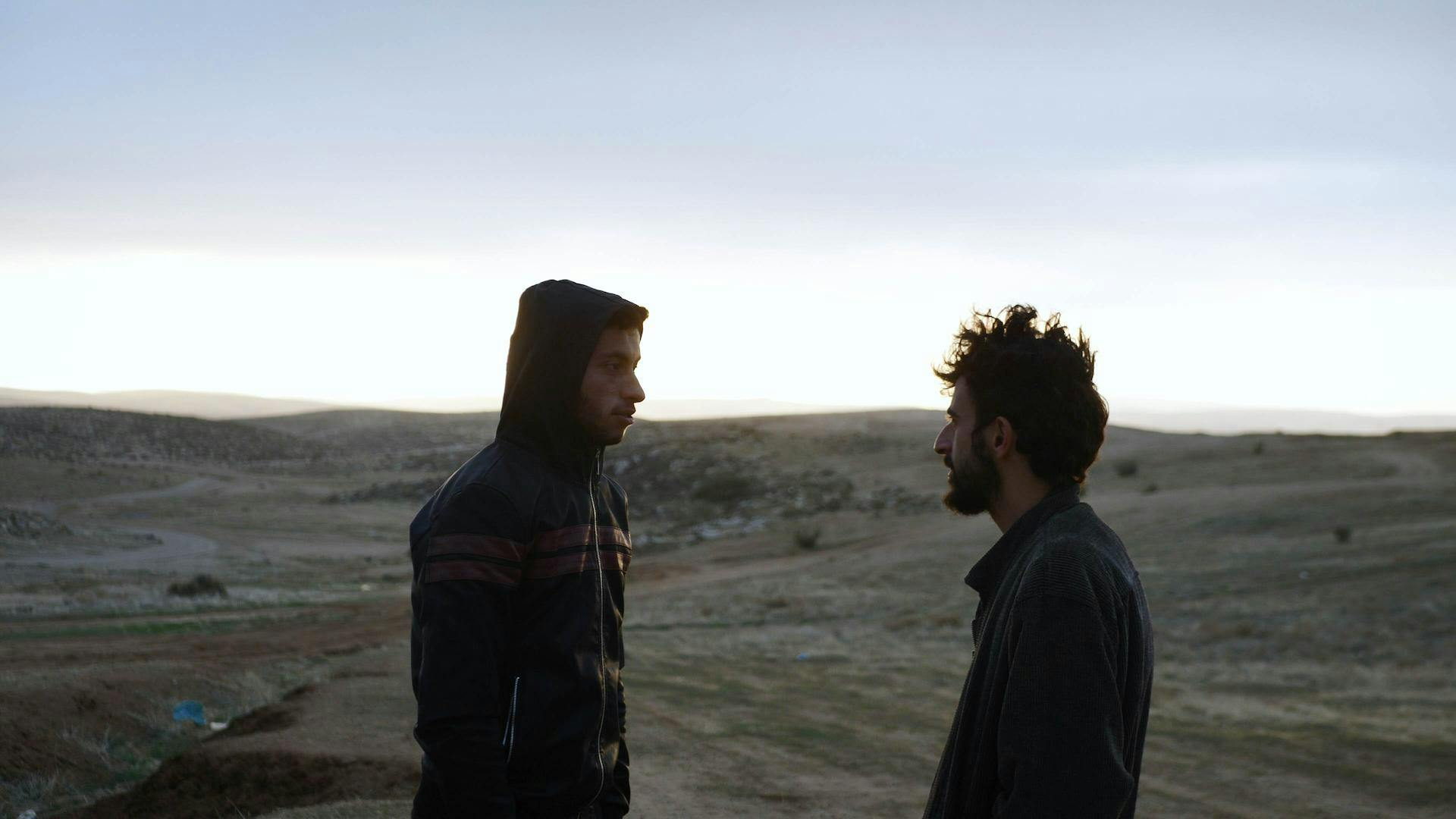 Photo from the film in which two people are looking at one another on a rolling hill landscape with a sunrise in the background