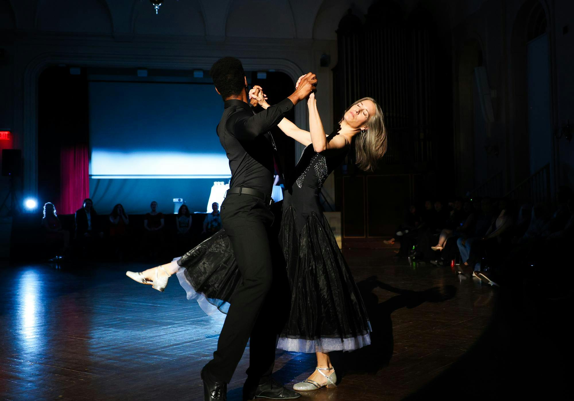 A man in all black dips a woman in a black dress in a ballroom.